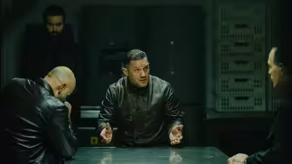 Harry Da Souza (Tom Hardy) gesturing while seated at a conference table