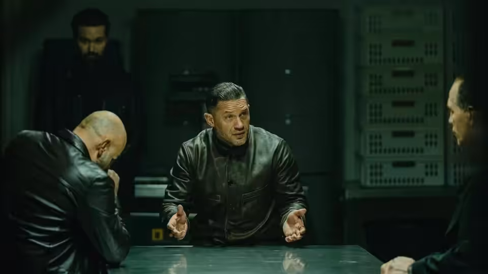 Harry Da Souza (Tom Hardy) gesturing while seated at a conference table