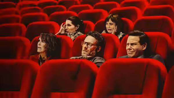 Seth Rogen sits with three others in a theater with red seats