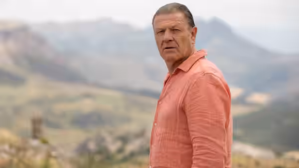 Man in orange shirt stands in mountainous landscape looking to one side