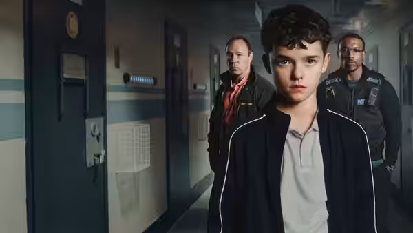 A boy in a tracksuit walking ahead of two men down an institutional corridor
