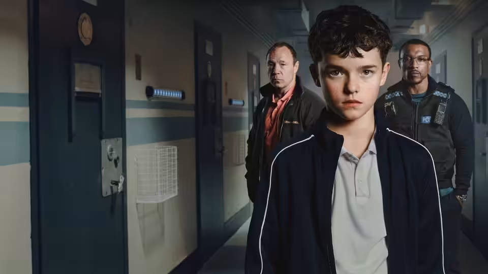 A boy in a tracksuit walking ahead of two men down an institutional corridor