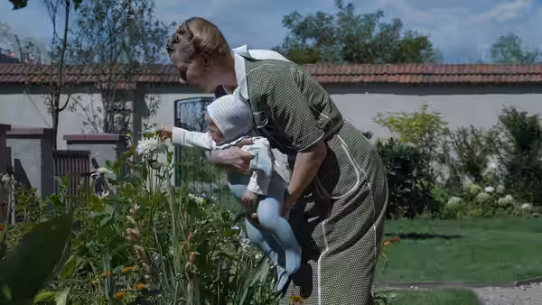 Woman holds infant while pointing to white flowers in garden