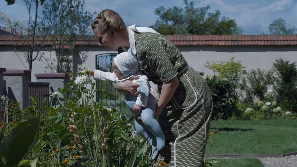 Woman holds infant while pointing to white flowers in garden