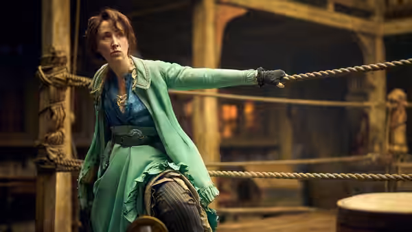 A woman in a green coat grips a rope while kneeling in a wooden boxing ring