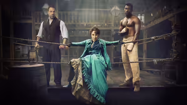 A woman in a teal dress stands in a boxing ring holding ropes flanked by two men