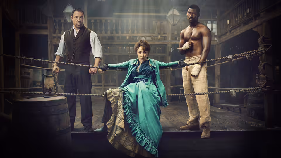 A woman in a teal dress stands in a boxing ring holding ropes flanked by two men
