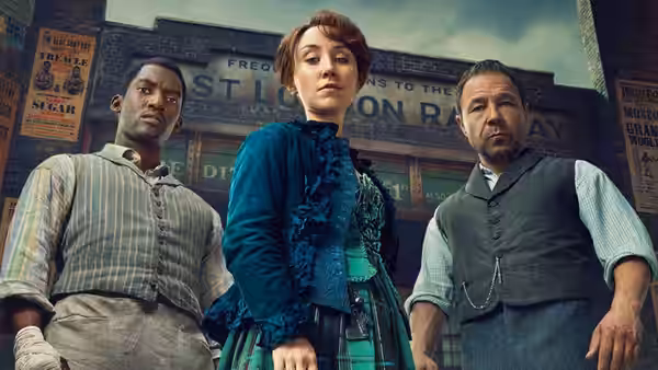 A woman in teal stands between two men in front of a railway station building