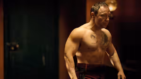 A muscular shirtless man with tattoos stands in a dimly lit room