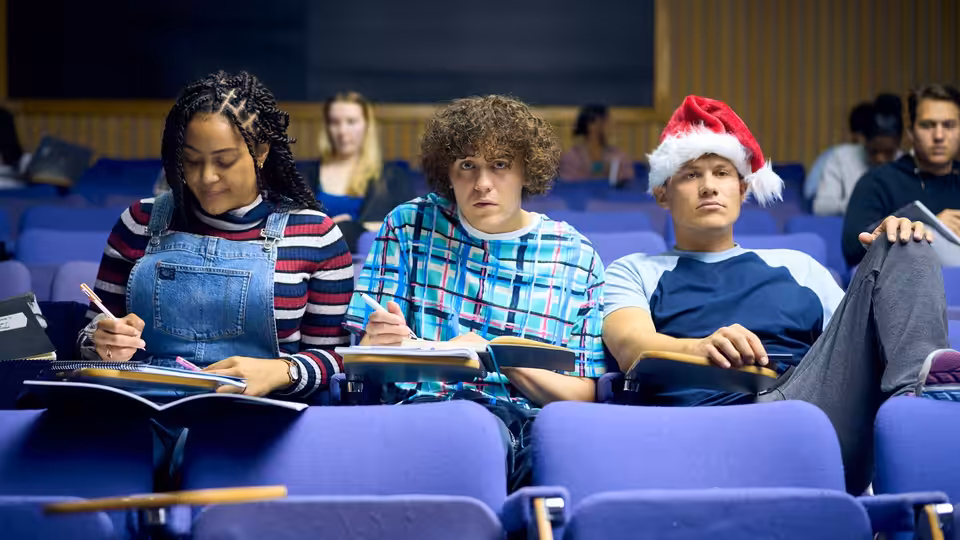 Three students sit in blue lecture hall seats with notebooks open