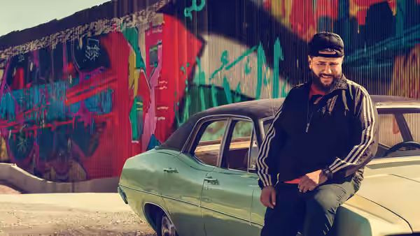 Mohammed Amer as Mo Najjar leaning against a green vintage car near a graffiti wall