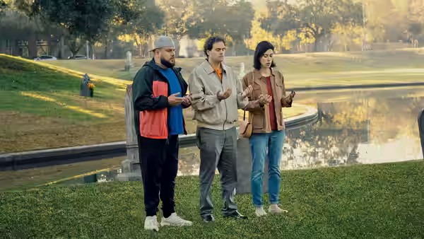 Mohammed Amer as Mo Najjar standing with two others by a pond in a park