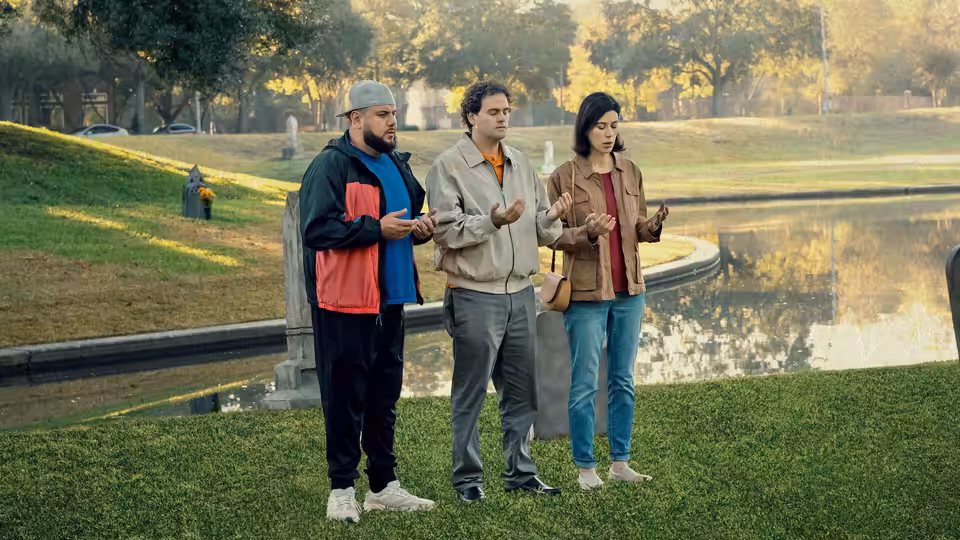 Mohammed Amer as Mo Najjar standing with two others by a pond in a park