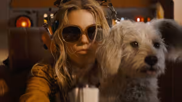 A blonde woman in sunglasses and headphones sitting beside a small white dog
