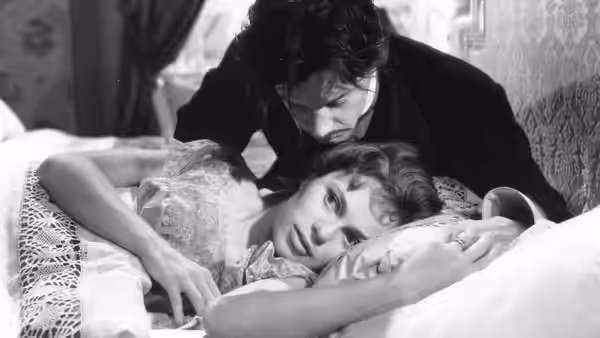 A young man leans over a woman lying in bed