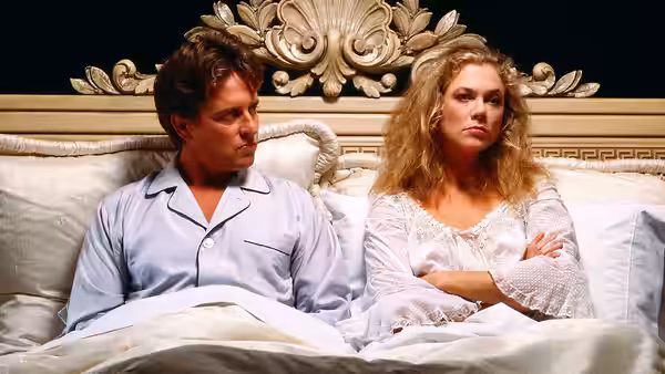 Oliver and Barbara sit apart in bed, arms crossed beneath an ornate headboard