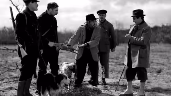 Several men with dogs and rifles gathered outdoors in a field during a discussion