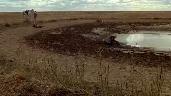 A person kneels beside a muddy pool as two figures stand near a horse in the distance