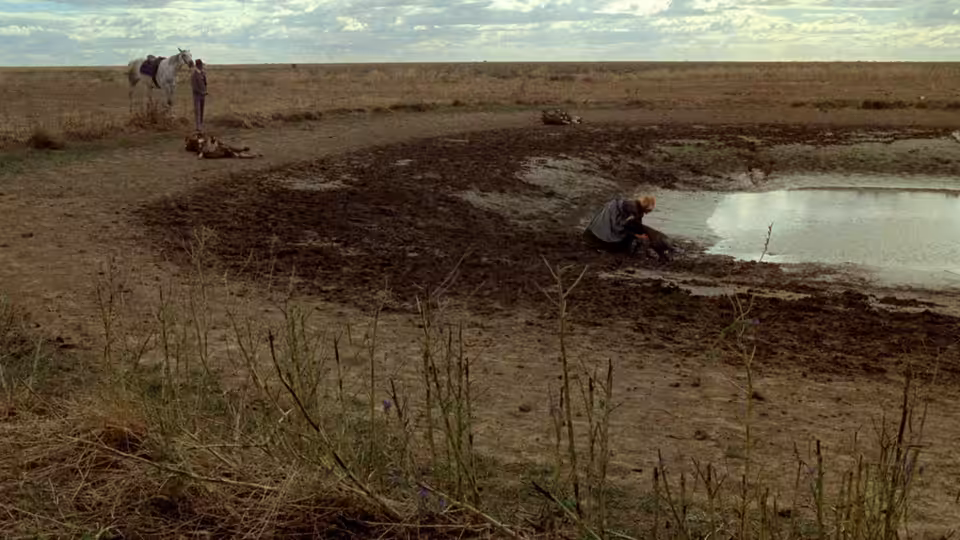 A person kneels beside a muddy pool as two figures stand near a horse in the distance