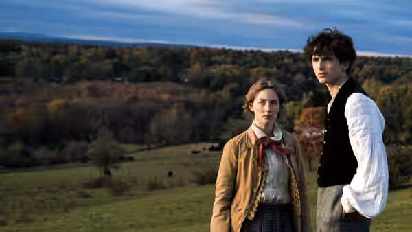 A young woman and a young man standing in a grassy field overlooking autumn hills