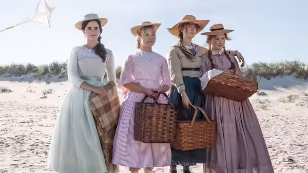Four women in Victorian dresses and hats standing on a beach holding picnic baskets