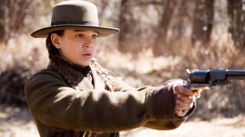 Mattie Ross (Hailee Steinfeld) aiming a revolver in the woods