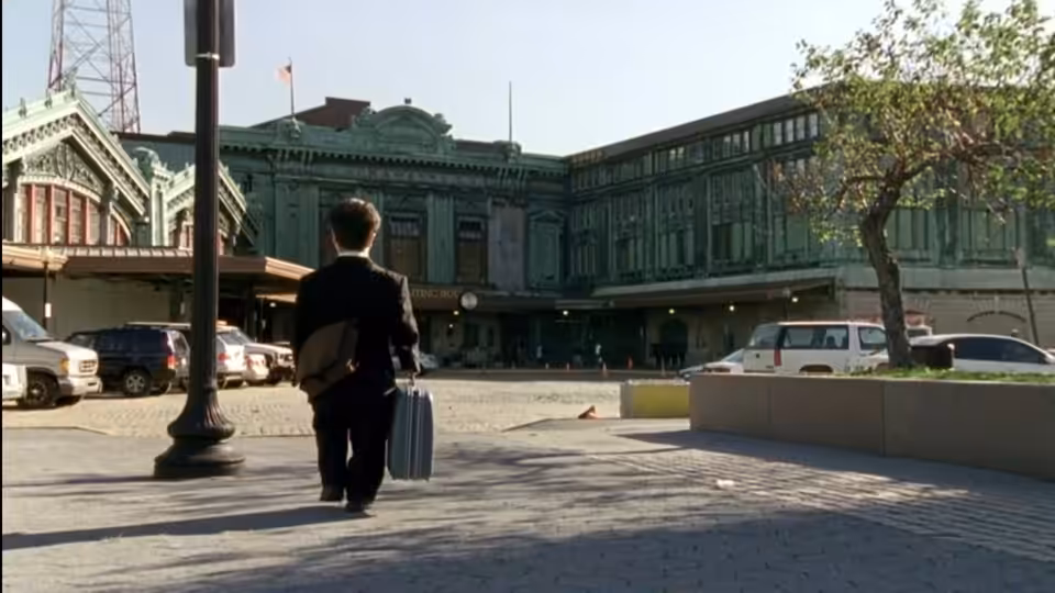 Finbar McBride (Peter Dinklage) carrying luggage toward a large green train station