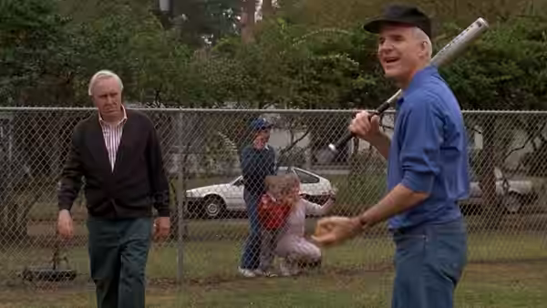 Gil Buckman (Steve Martin) holds a baseball bat near a fenced yard as Frank Buckman approaches
