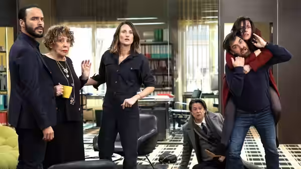 In a bright office a woman gestures while a man carries another on his back