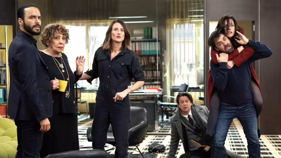 In a bright office a woman gestures while a man carries another on his back