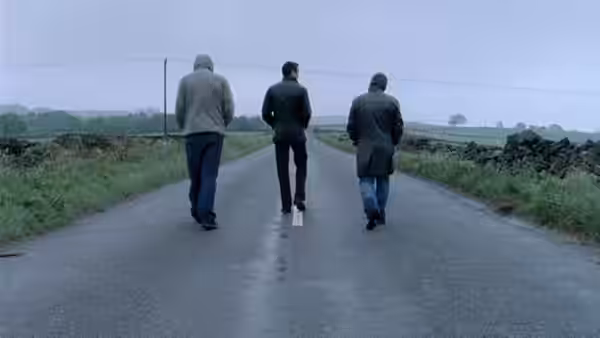 Three men walk away along a wet rural road under an overcast sky