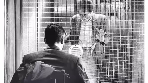 A man behind wire mesh gesturing toward another seated man