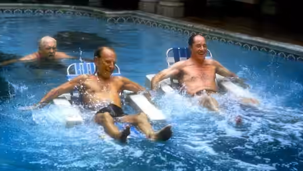 Three elderly men splash in a swimming pool while seated in lounge chairs