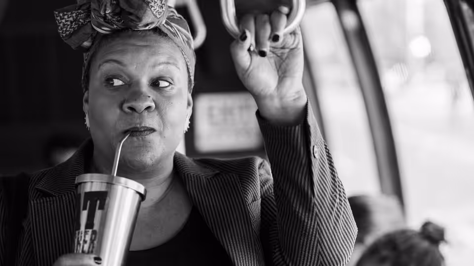 Radha (Radha Blank) sips from a cup while gripping an overhead strap on public transit