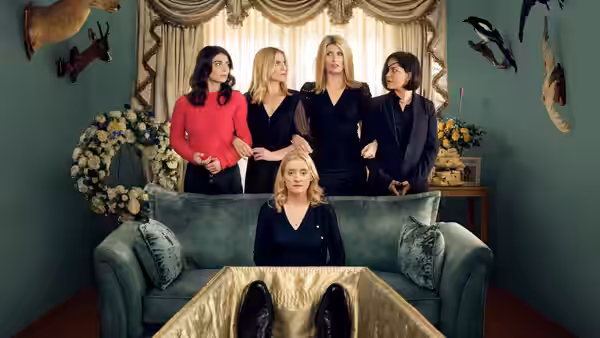 five women gathered around a green couch with a golden coffin in front