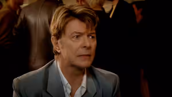 David Bowie in a grey suit looks to his left with neutral expression