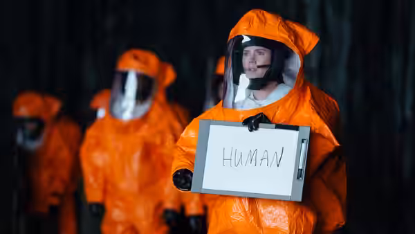 A man in an orange hazmat suit holds a whiteboard labeled HUMAN