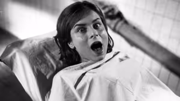 A dark-haired woman lies on an operating table, staring wide-eyed with her mouth open
