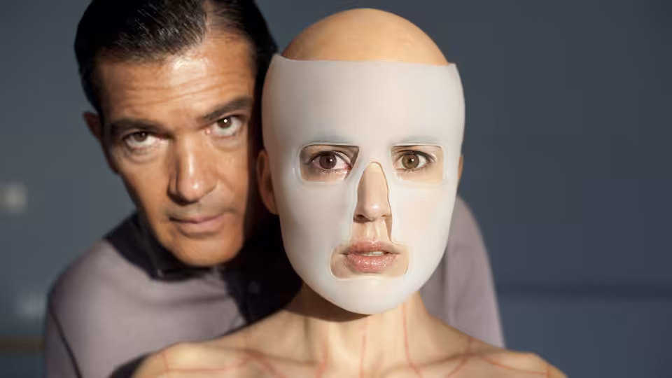 Dr. Robert Ledgard (Antonio Banderas) stands behind a woman wearing a pale face mask