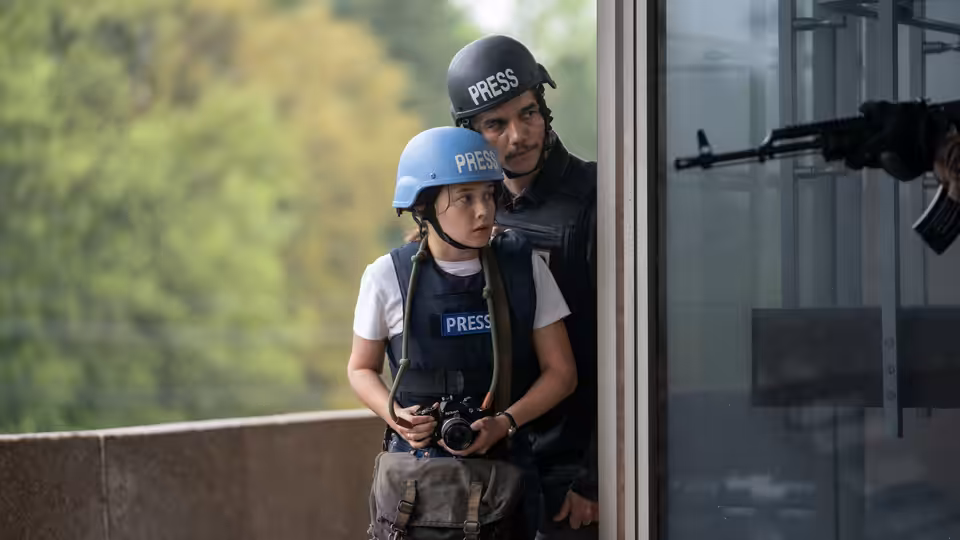 Jessie (Cailee Spaeny) and Joel (Wagner Moura) in press vests peering from a window