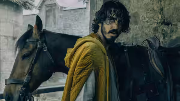 Gawain (Dev Patel) stands beside his saddled horse in a stone courtyard