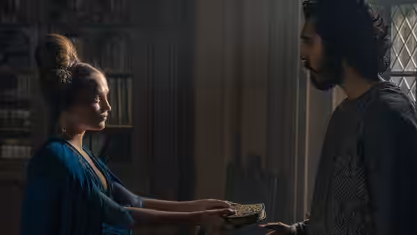 Lady (Alicia Vikander) offers Gawain (Dev Patel) a small object in a book-lined room