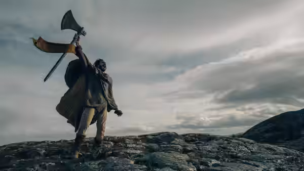 Gawain (Dev Patel) raises an axe atop a rocky ridge beneath a cloudy sky