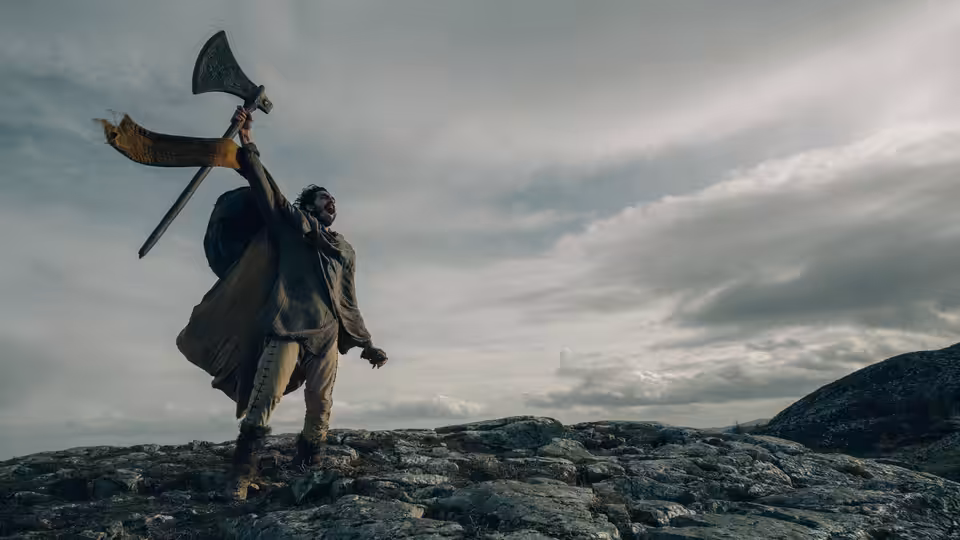 Gawain (Dev Patel) raises an axe atop a rocky ridge beneath a cloudy sky