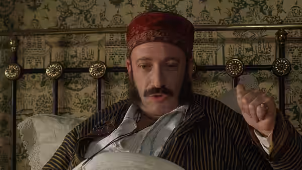 Man in striped pajamas and red cap sits up in bed gesturing with one hand