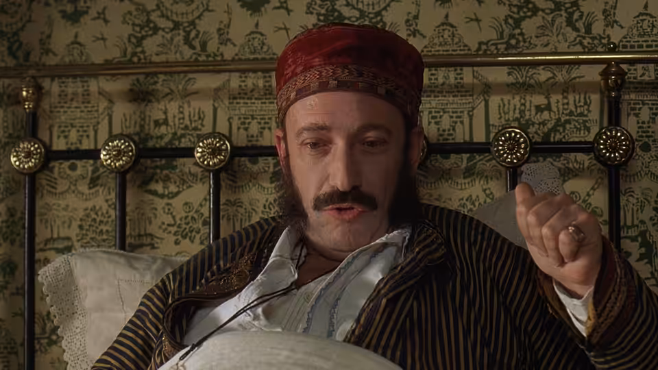 Man in striped pajamas and red cap sits up in bed gesturing with one hand