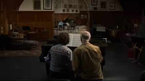 two men seated at a grand piano studying sheet music in a rehearsal room