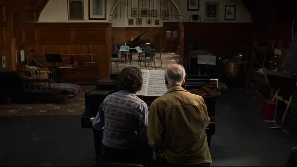 two men seated at a grand piano studying sheet music in a rehearsal room