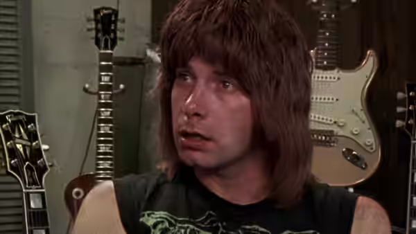 Man with shaggy brown hair sits among electric guitars in a room