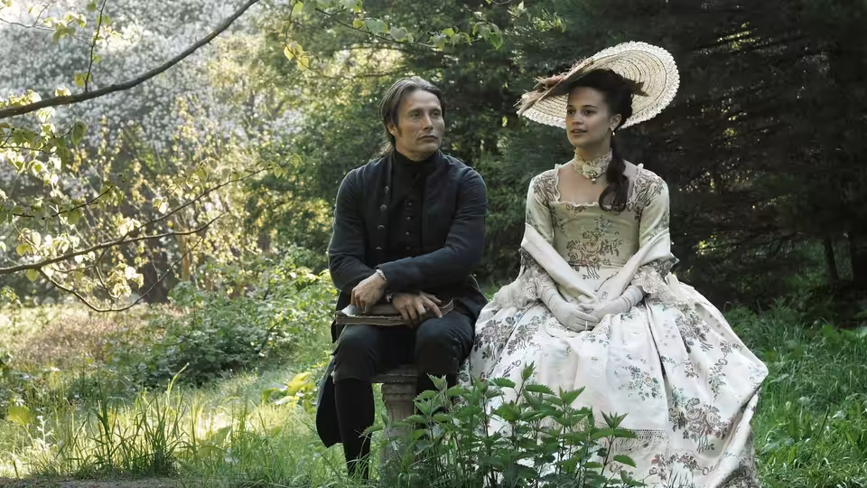 Johann Friedrich Struensee (Mads Mikkelsen) sits beside Caroline Mathilde (Alicia Vikander) in a garden